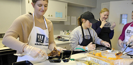 Four girls wearing aprons and hairnets scoop food into trays