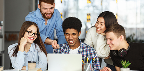 Young adults sit and stand behind computer screen smiling.